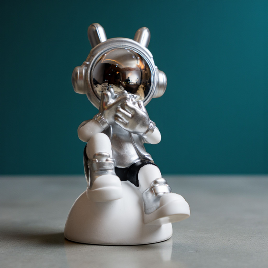 Seated astronaut figurine in metallic silver and white covering its visor in a speak-no-evil pose