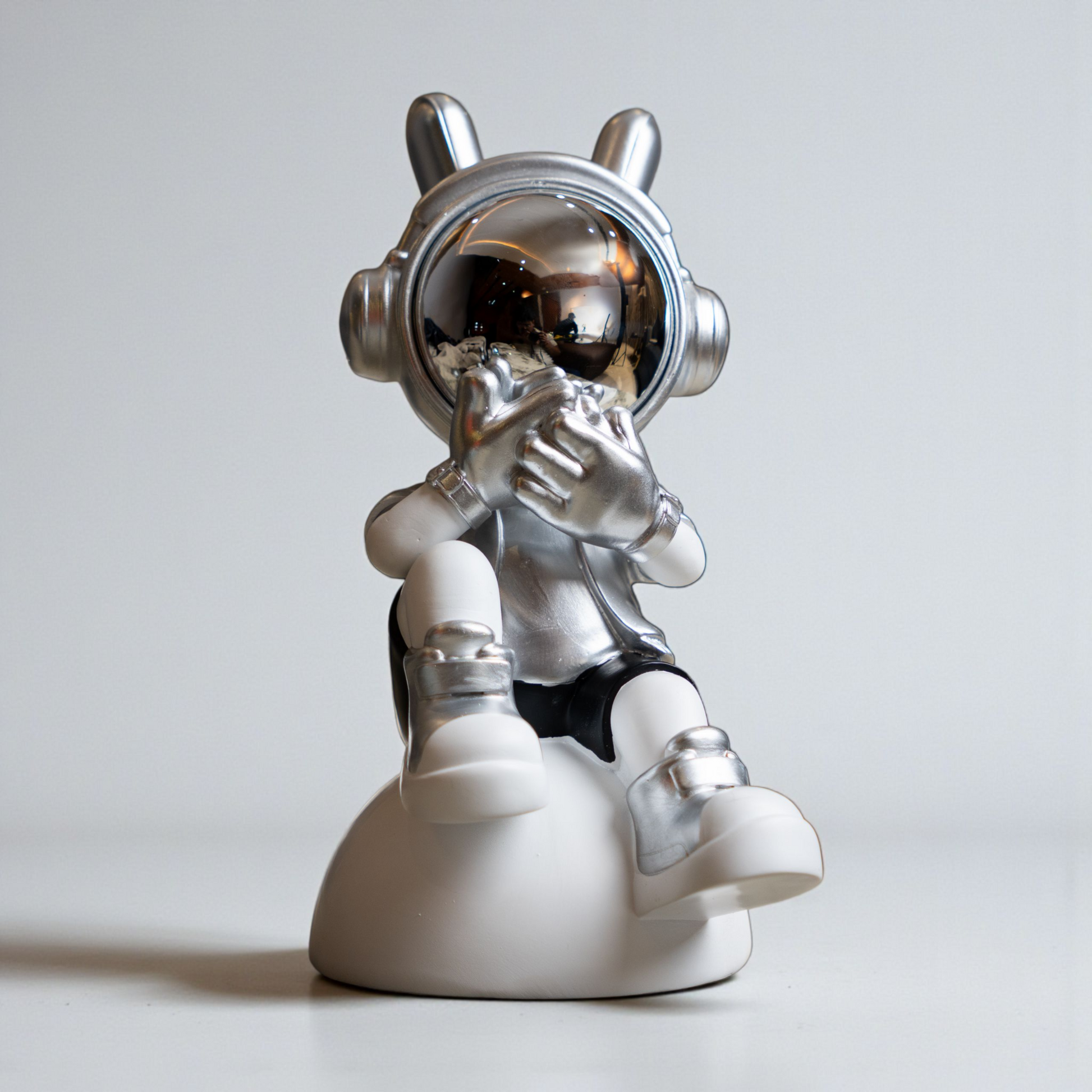 Seated astronaut figurine in metallic silver and white covering its visor in a speak-no-evil pose