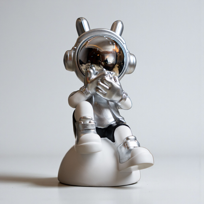 Seated astronaut figurine in metallic silver and white covering its visor in a speak-no-evil pose