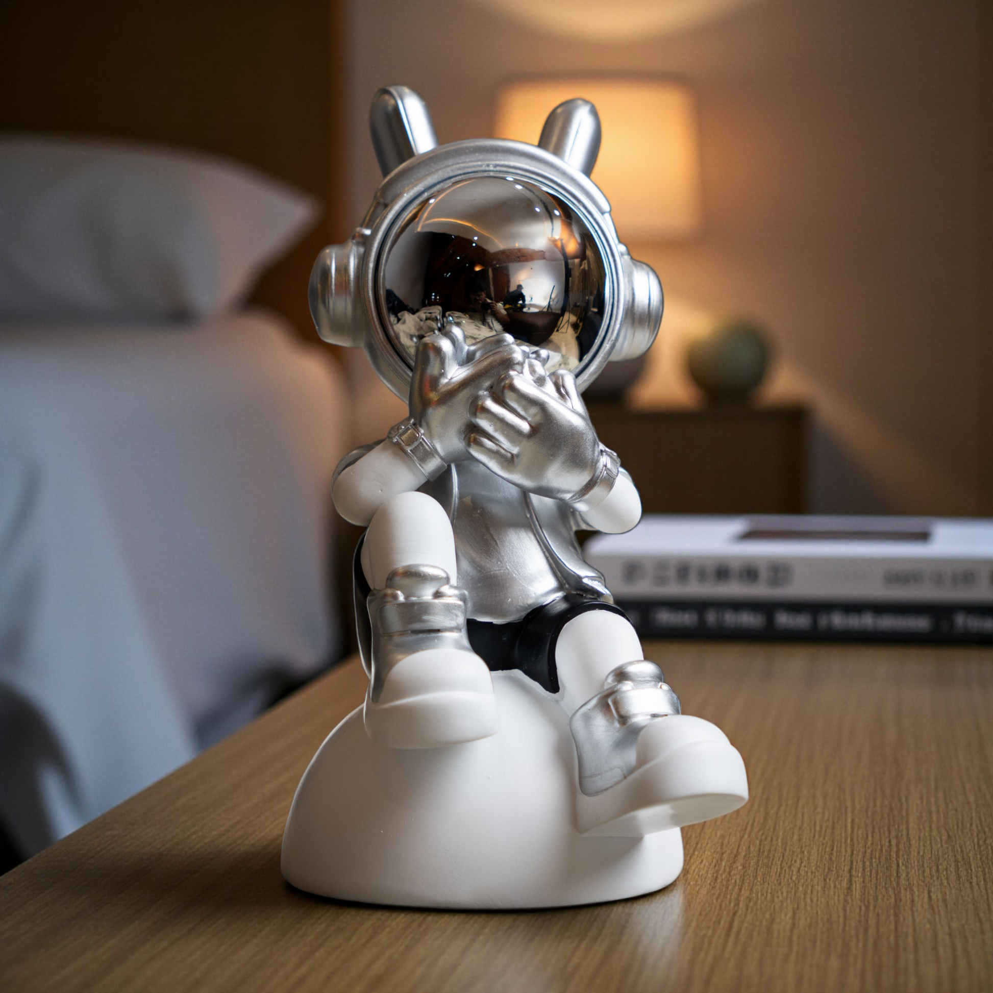 Seated astronaut figurine in metallic silver and white covering its visor in a speak-no-evil pose