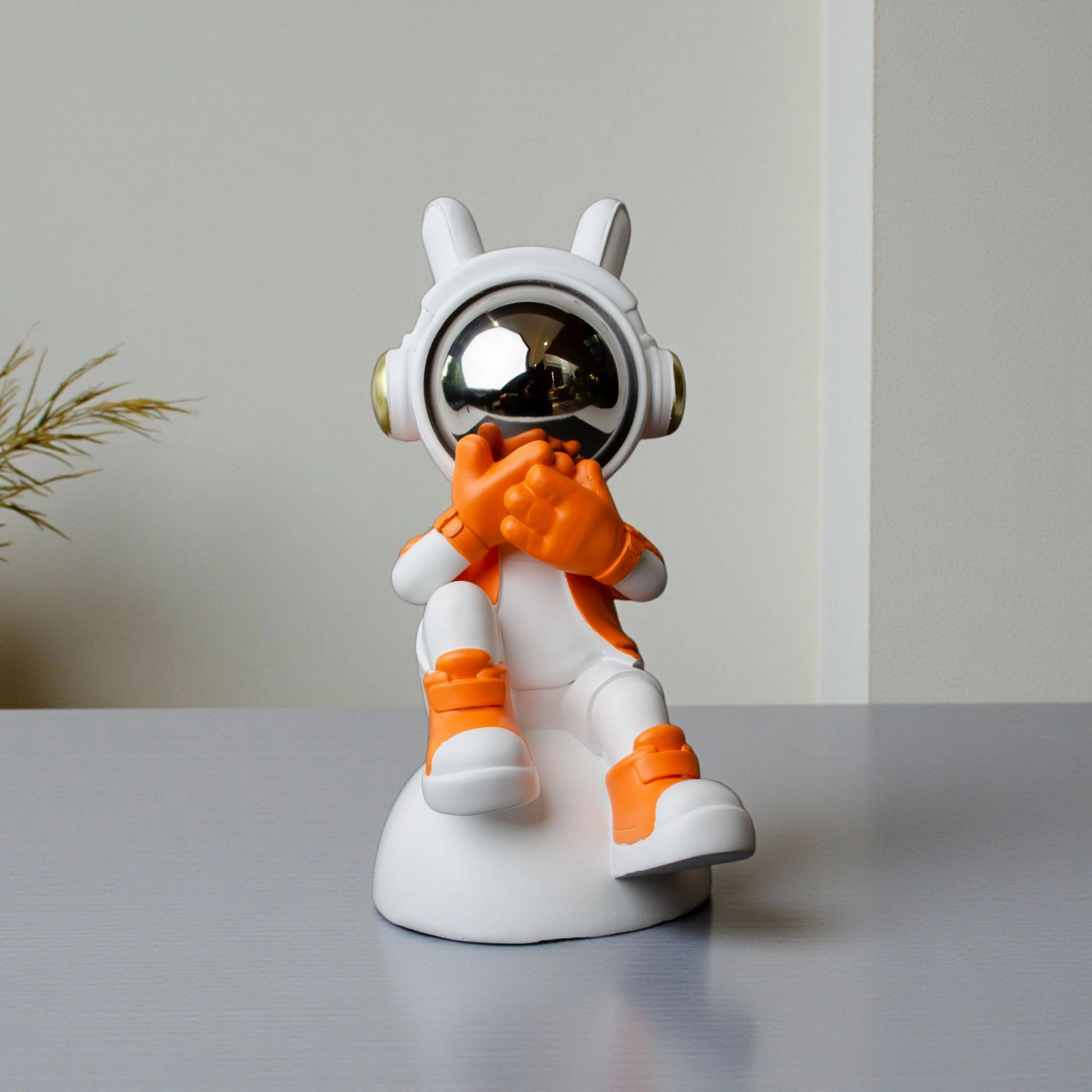Astronaut whisper figurine in white and orange with chrome visor seated on a base—modern resin décor showpiece