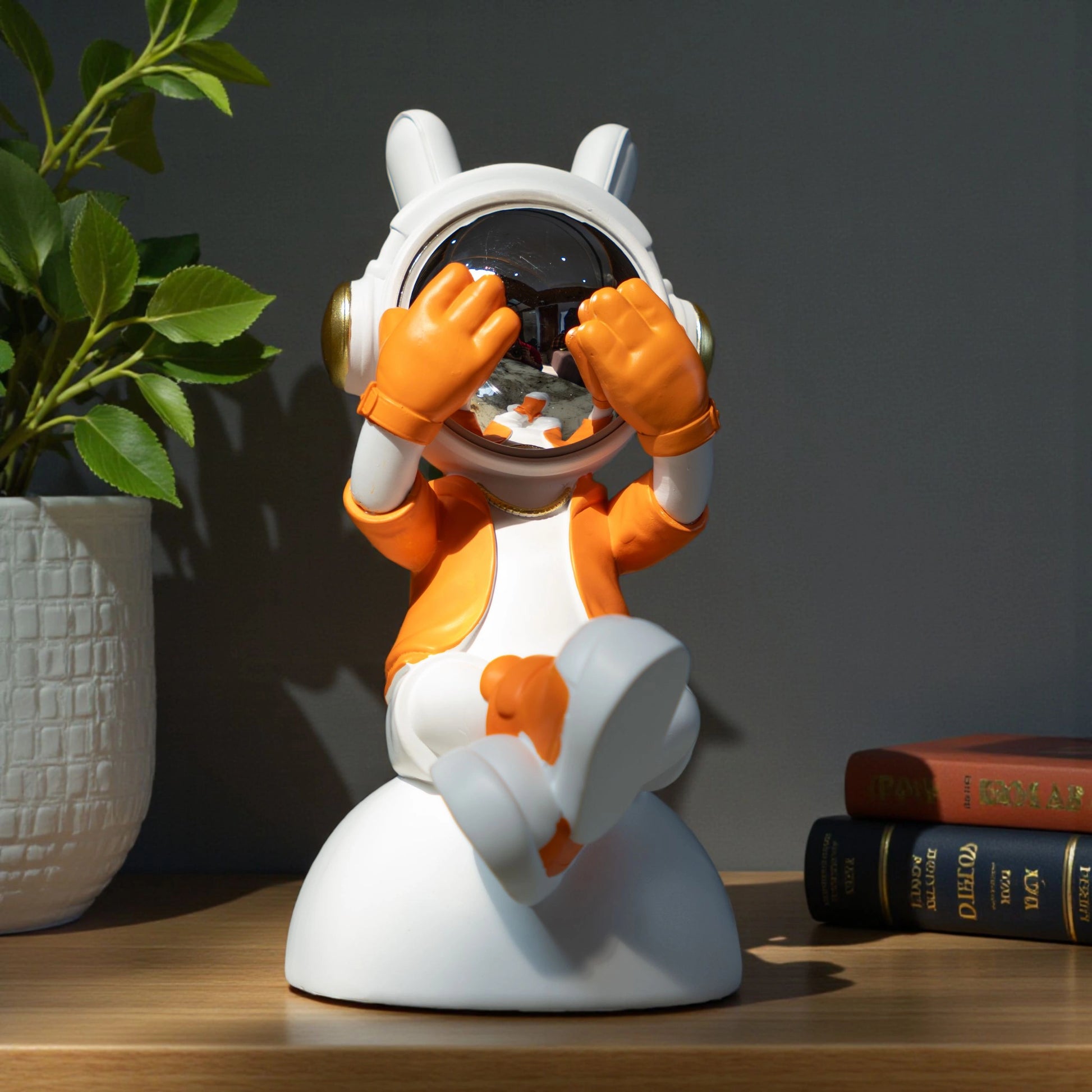Cute astronaut figurine in orange and white with chrome visor covering its eyes in a playful peek pose, seated on a rounded base; modern resin décor showpiece