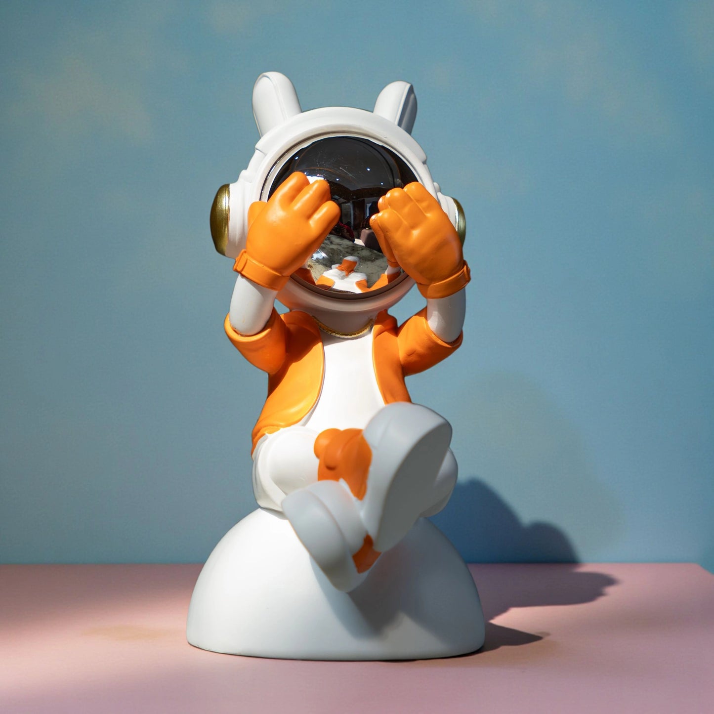 Cute astronaut figurine in orange and white with chrome visor covering its eyes in a playful peek pose, seated on a rounded base; modern resin décor showpiece
