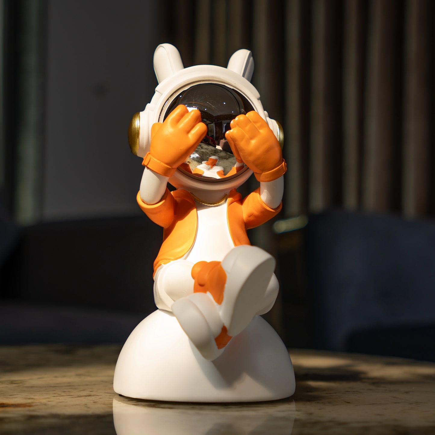 Cute astronaut figurine in orange and white with chrome visor covering its eyes in a playful peek pose, seated on a rounded base; modern resin décor showpiece