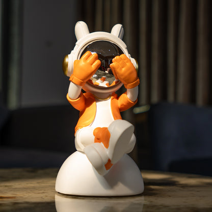 Cute astronaut figurine in orange and white with chrome visor covering its eyes in a playful peek pose, seated on a rounded base; modern resin décor showpiece