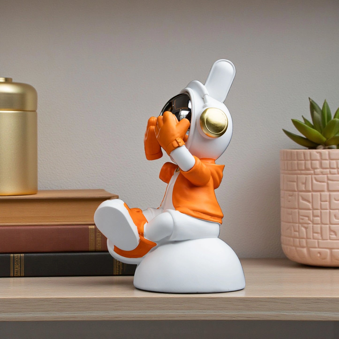 Cute astronaut figurine in orange and white with chrome visor covering its eyes in a playful peek pose, seated on a rounded base; modern resin décor showpiece