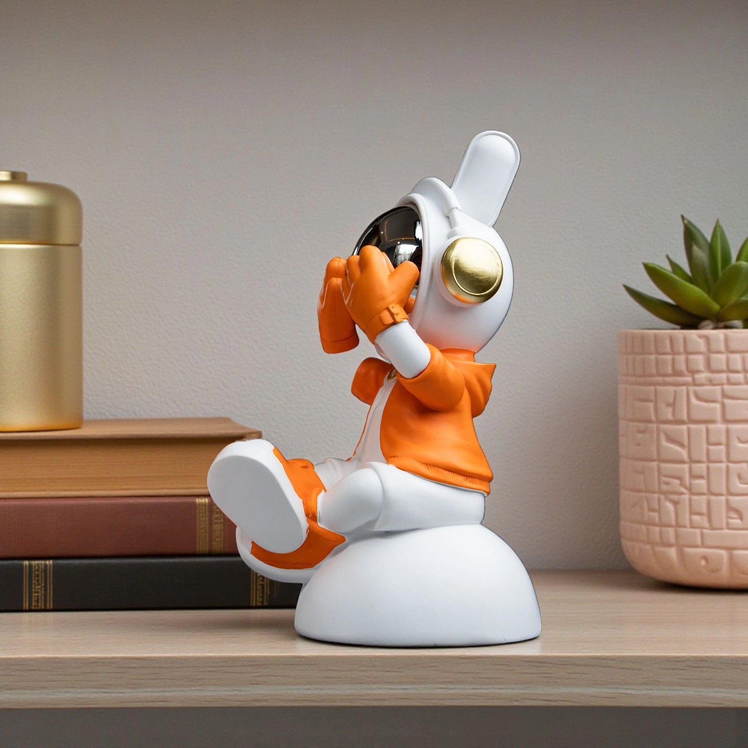 Cute astronaut figurine in orange and white with chrome visor covering its eyes in a playful peek pose, seated on a rounded base; modern resin décor showpiece