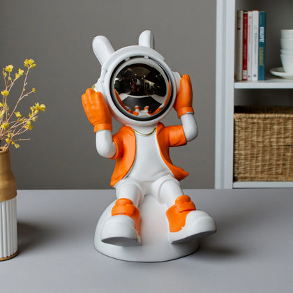 Astronaut figurine in orange and white holding its helmet sides in a listening pose with a chrome visor, seated on a dome-shaped base; modern resin décor piece