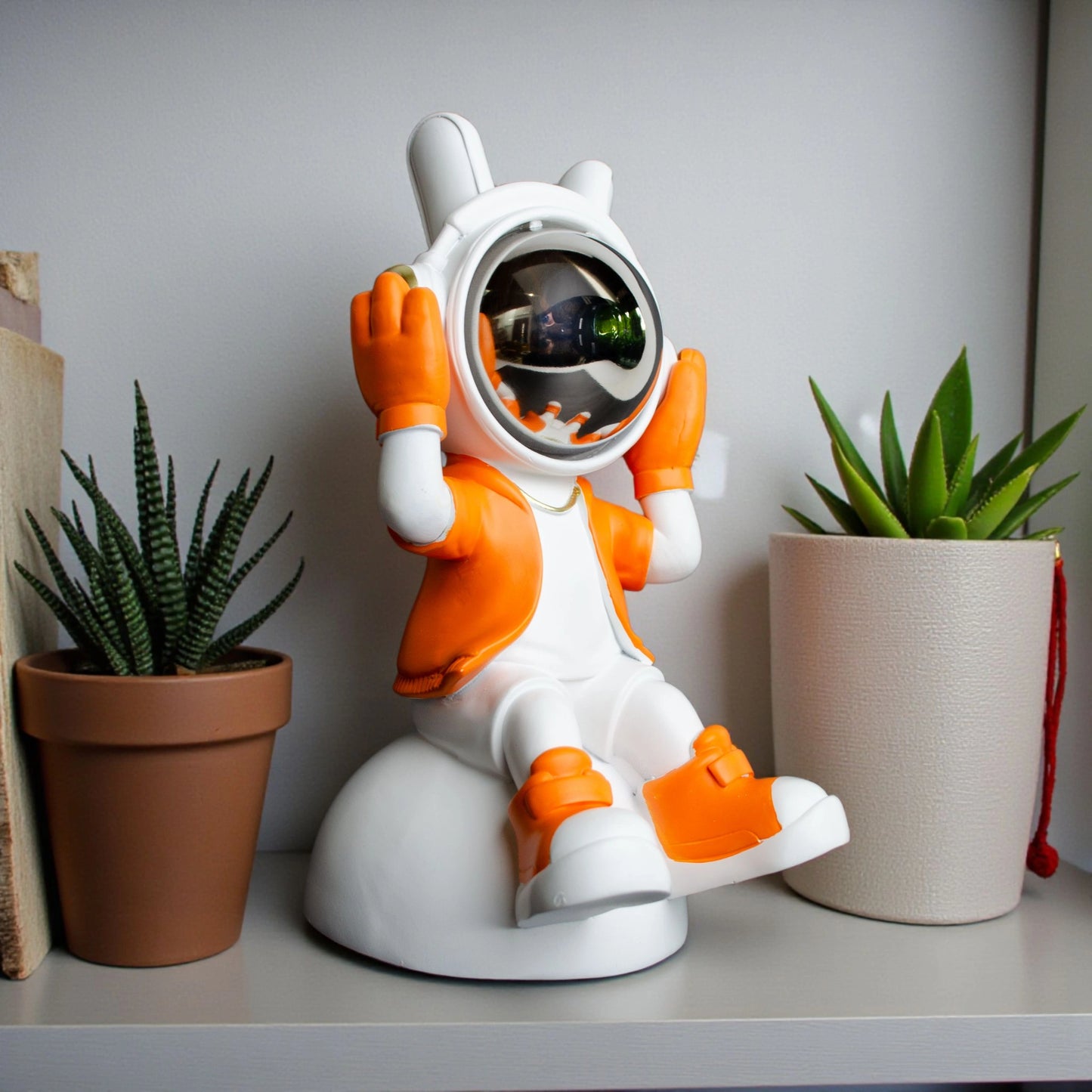Astronaut figurine in orange and white holding its helmet sides in a listening pose with a chrome visor, seated on a dome-shaped base; modern resin décor piece