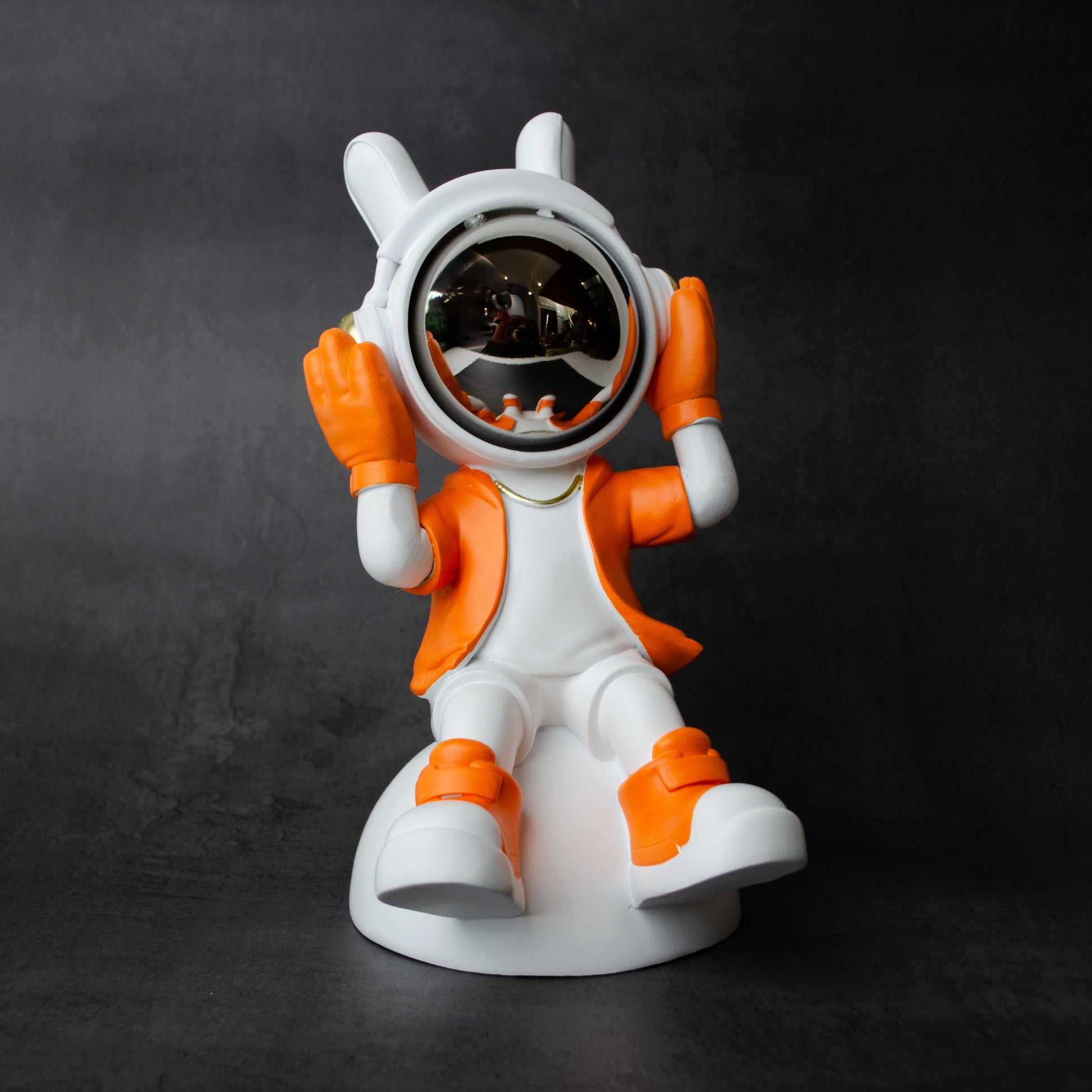 Astronaut figurine in orange and white holding its helmet sides in a listening pose with a chrome visor, seated on a dome-shaped base; modern resin décor piece