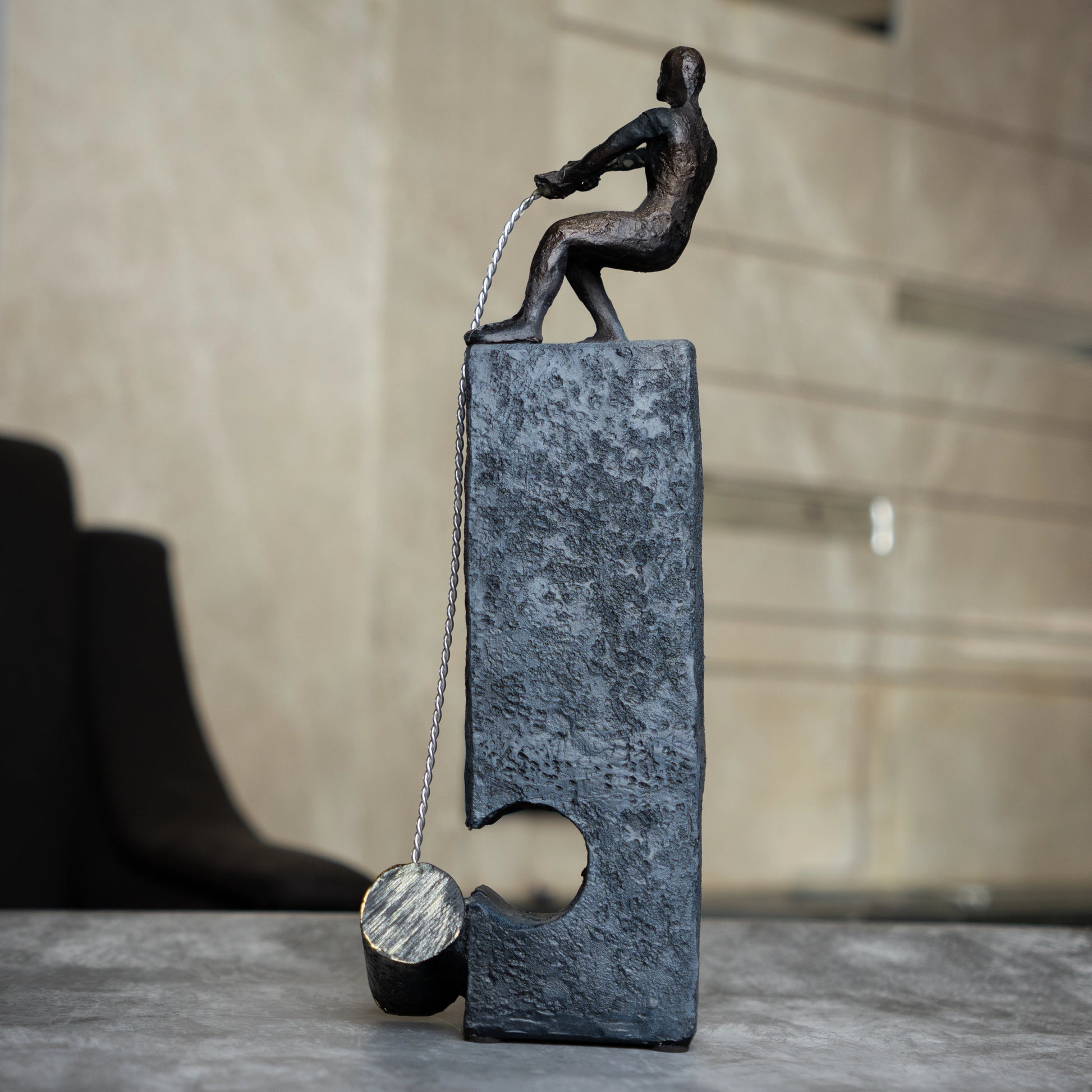 Bronze pulling figure sculpture on textured charcoal stone base with hanging weight.