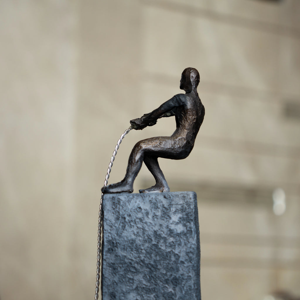 Bronze pulling figure sculpture on textured charcoal stone base with hanging weight.