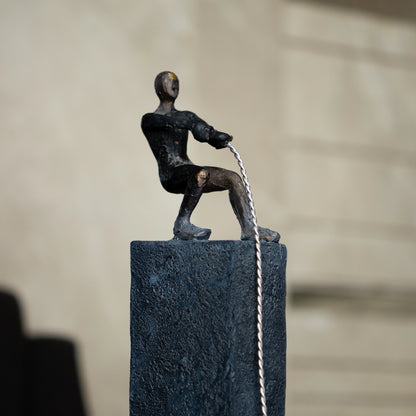 Bronze pulling figure sculpture on textured charcoal stone base with hanging weight.