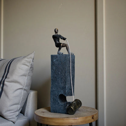 Bronze pulling figure sculpture on textured charcoal stone base with hanging weight.
