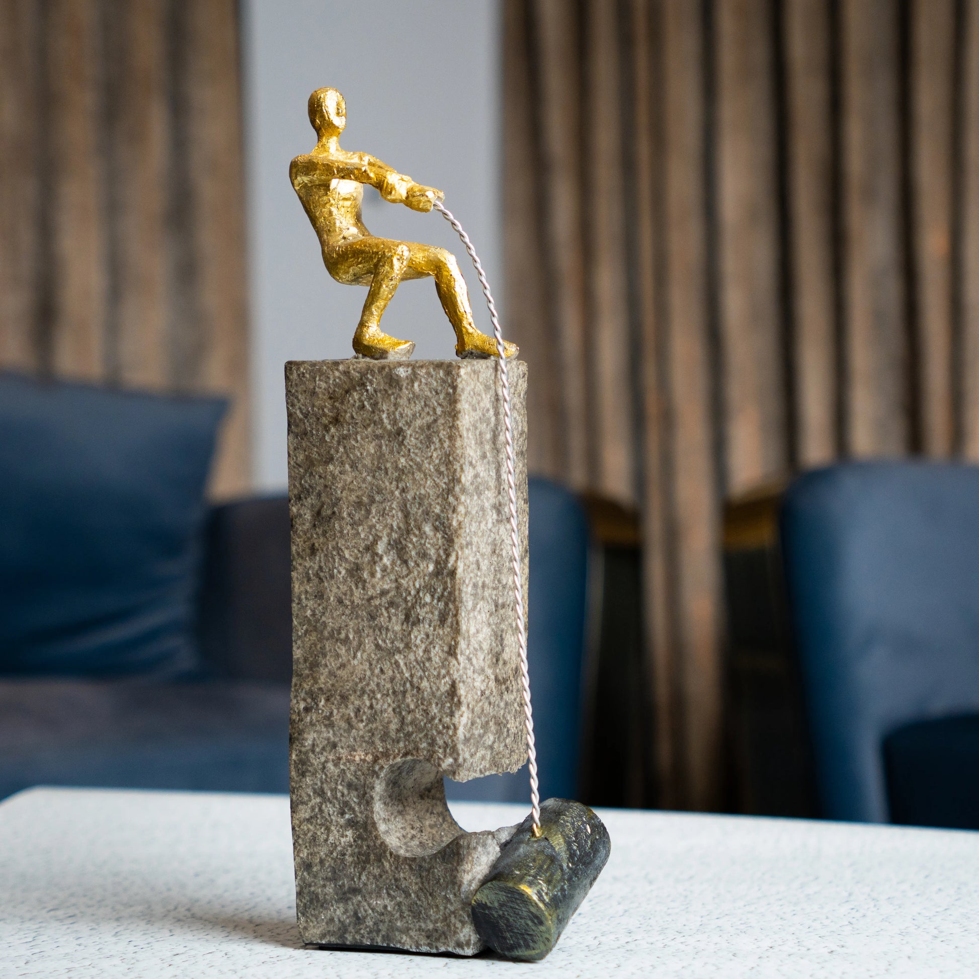 Gold pulling figure sculpture on textured charcoal stone base with hanging weight.
