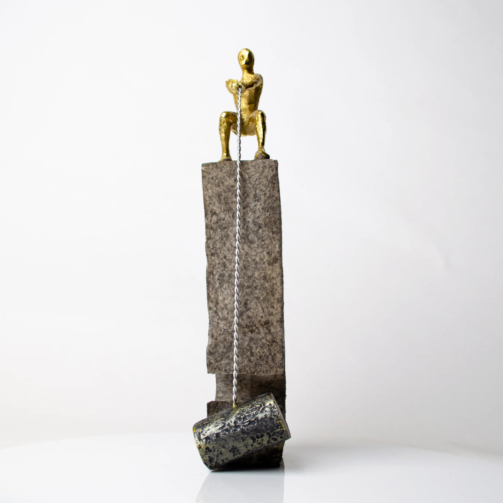 Gold pulling figure sculpture on textured charcoal stone base with hanging weight.