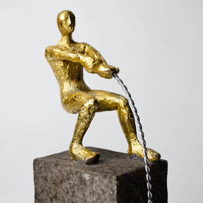 Gold pulling figure sculpture on textured charcoal stone base with hanging weight.