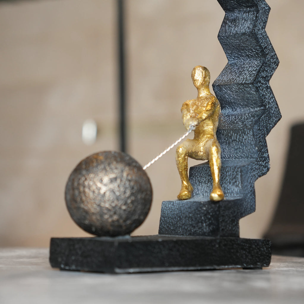 Modern abstract human figure decor seated on geometric staircase pulling a sphere, black and gold tabletop accent for living room, office desk or console styling