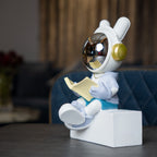 Astronaut figurine with chrome visor sitting cross-legged on a block while reading a book, wearing a white suit, gold accents, and blue pants; modern resin décor showpiece