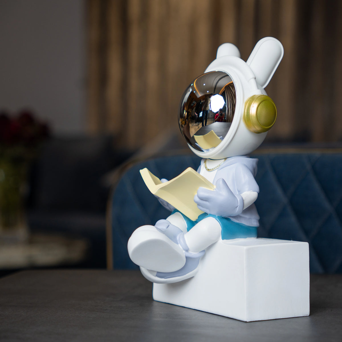 Astronaut figurine with chrome visor sitting cross-legged on a block while reading a book, wearing a white suit, gold accents, and blue pants; modern resin décor showpiece