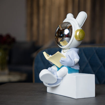 Astronaut figurine with chrome visor sitting cross-legged on a block while reading a book, wearing a white suit, gold accents, and blue pants; modern resin décor showpiece