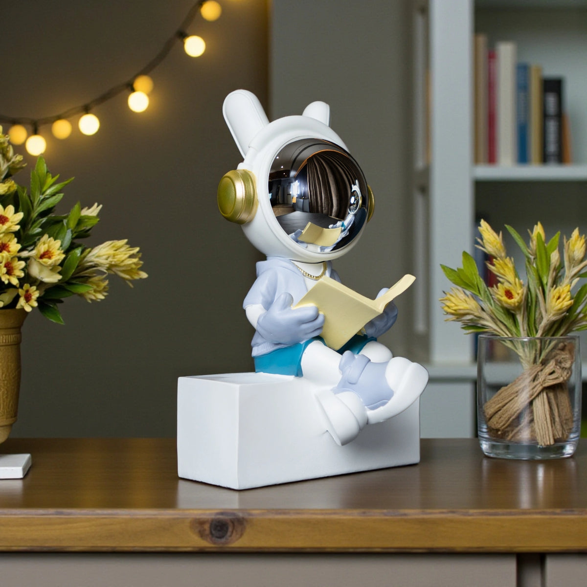 Astronaut figurine with chrome visor sitting cross-legged on a block while reading a book, wearing a white suit, gold accents, and blue pants; modern resin décor showpiece