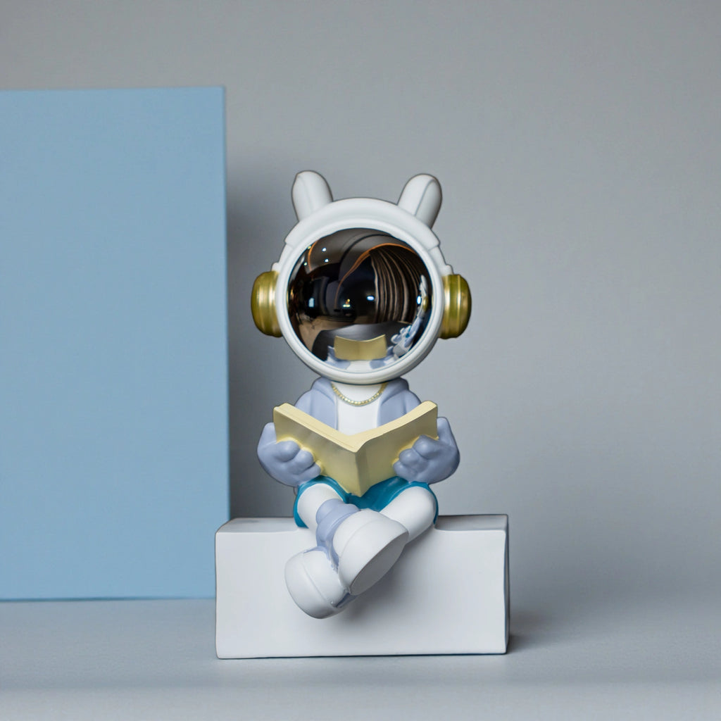 Astronaut figurine with chrome visor sitting cross-legged on a block while reading a book, wearing a white suit, gold accents, and blue pants; modern resin décor showpiece