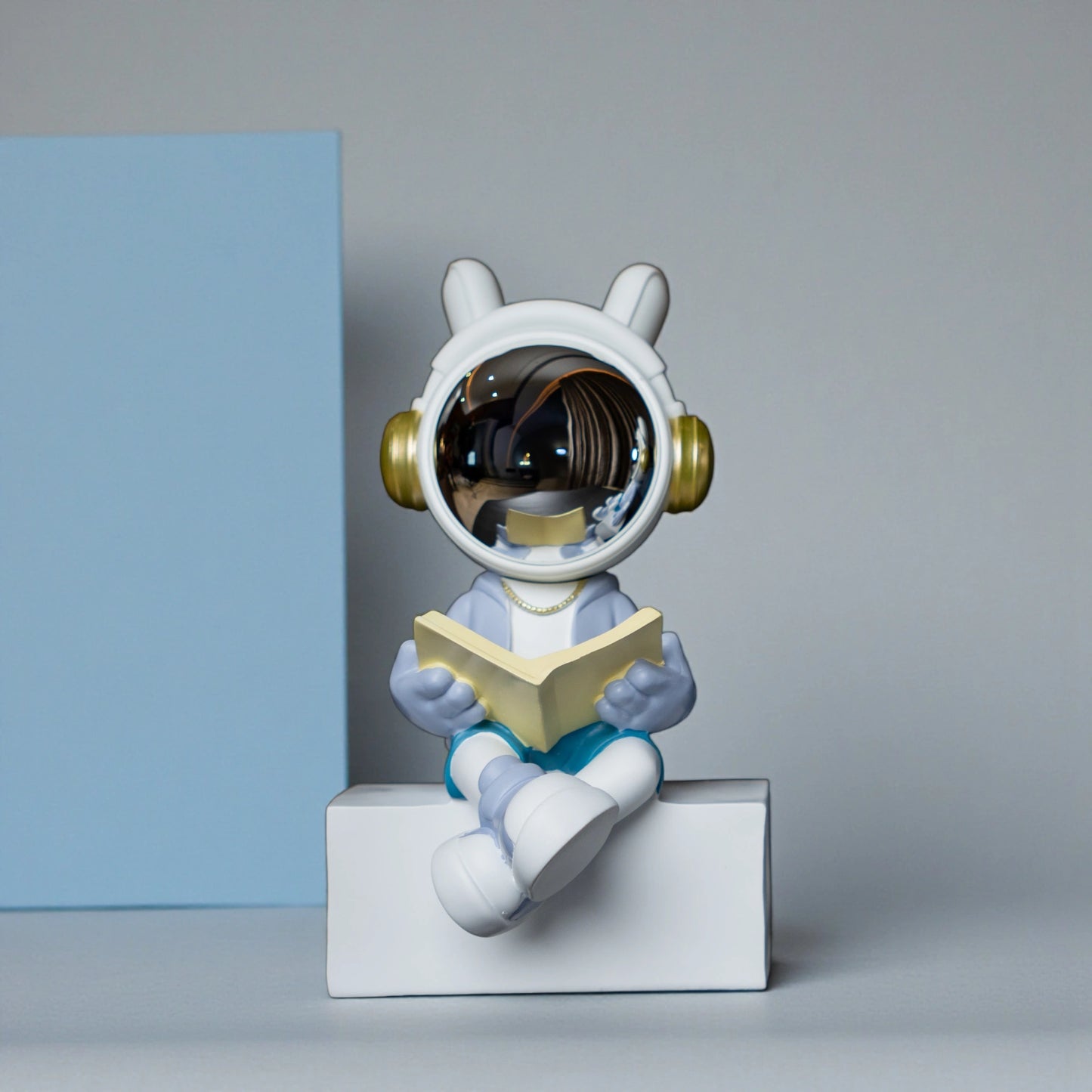 Astronaut figurine with chrome visor sitting cross-legged on a block while reading a book, wearing a white suit, gold accents, and blue pants; modern resin décor showpiece
