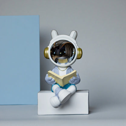 Astronaut figurine with chrome visor sitting cross-legged on a block while reading a book, wearing a white suit, gold accents, and blue pants; modern resin décor showpiece