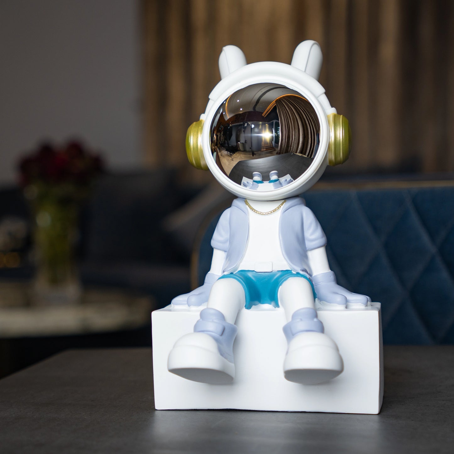 Cute astronaut figurine in blue and white with gold accents and a chrome visor sitting casually on a white block; modern resin décor piece
