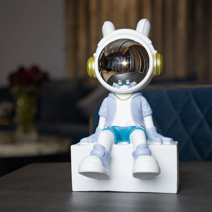 Cute astronaut figurine in blue and white with gold accents and a chrome visor sitting casually on a white block; modern resin décor piece