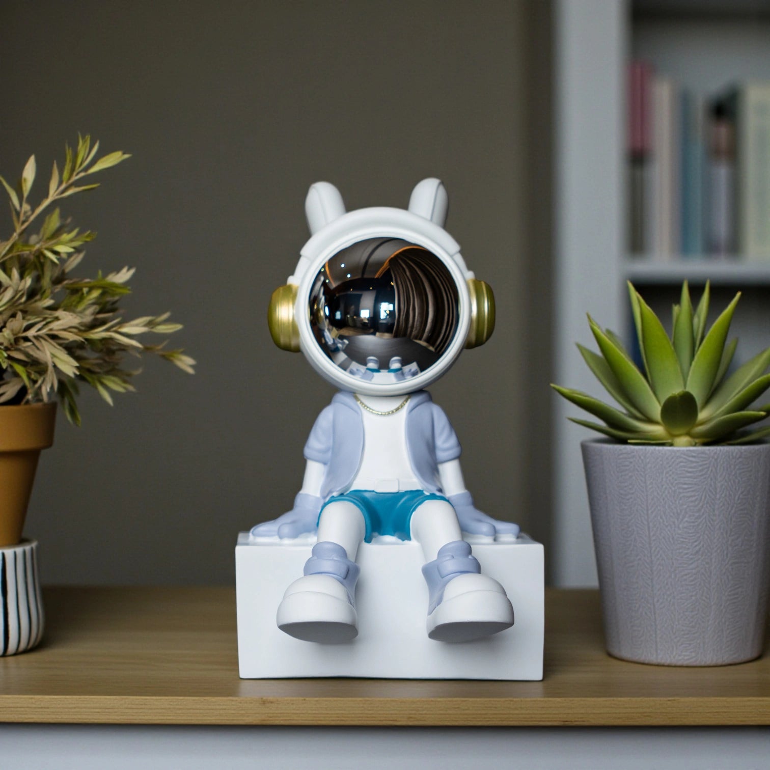 Cute astronaut figurine in blue and white with gold accents and a chrome visor sitting casually on a white block; modern resin décor piece