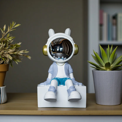 Cute astronaut figurine in blue and white with gold accents and a chrome visor sitting casually on a white block; modern resin décor piece
