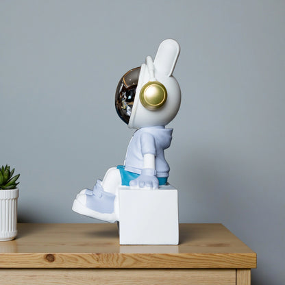 Cute astronaut figurine in blue and white with gold accents and a chrome visor sitting casually on a white block; modern resin décor piece