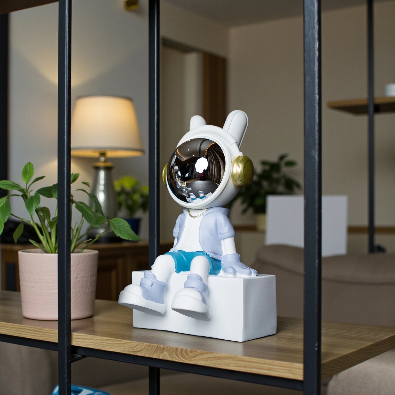 Cute astronaut figurine in blue and white with gold accents and a chrome visor sitting casually on a white block; modern resin décor piece