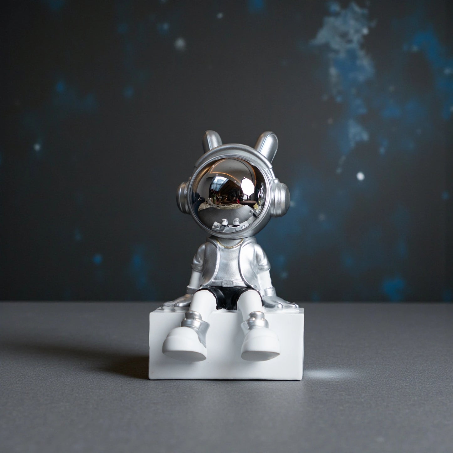 Silver astronaut figurine with chrome visor sitting casually on a white block, wearing metallic silver suit and black shorts; modern resin décor showpiece