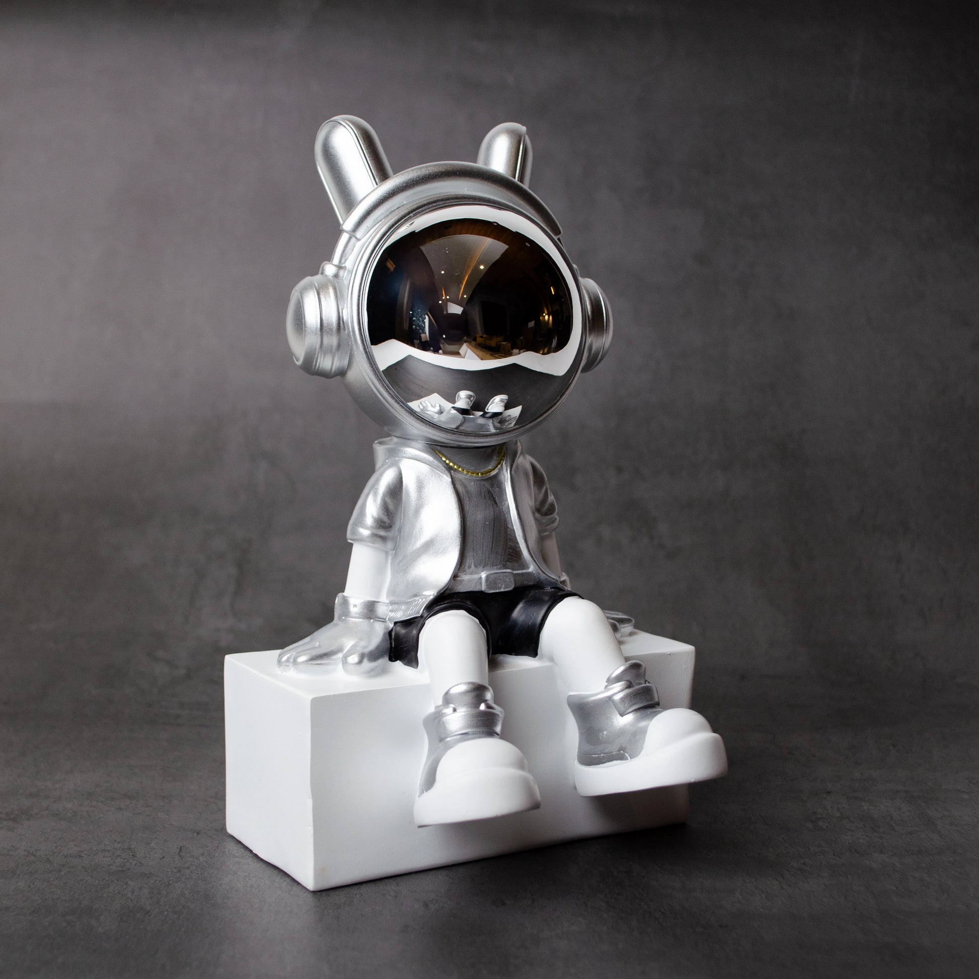 Silver astronaut figurine with chrome visor sitting casually on a white block, wearing metallic silver suit and black shorts; modern resin décor showpiece