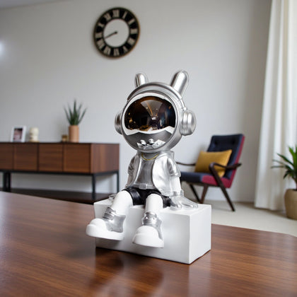 Silver astronaut figurine with chrome visor sitting casually on a white block, wearing metallic silver suit and black shorts; modern resin décor showpiece