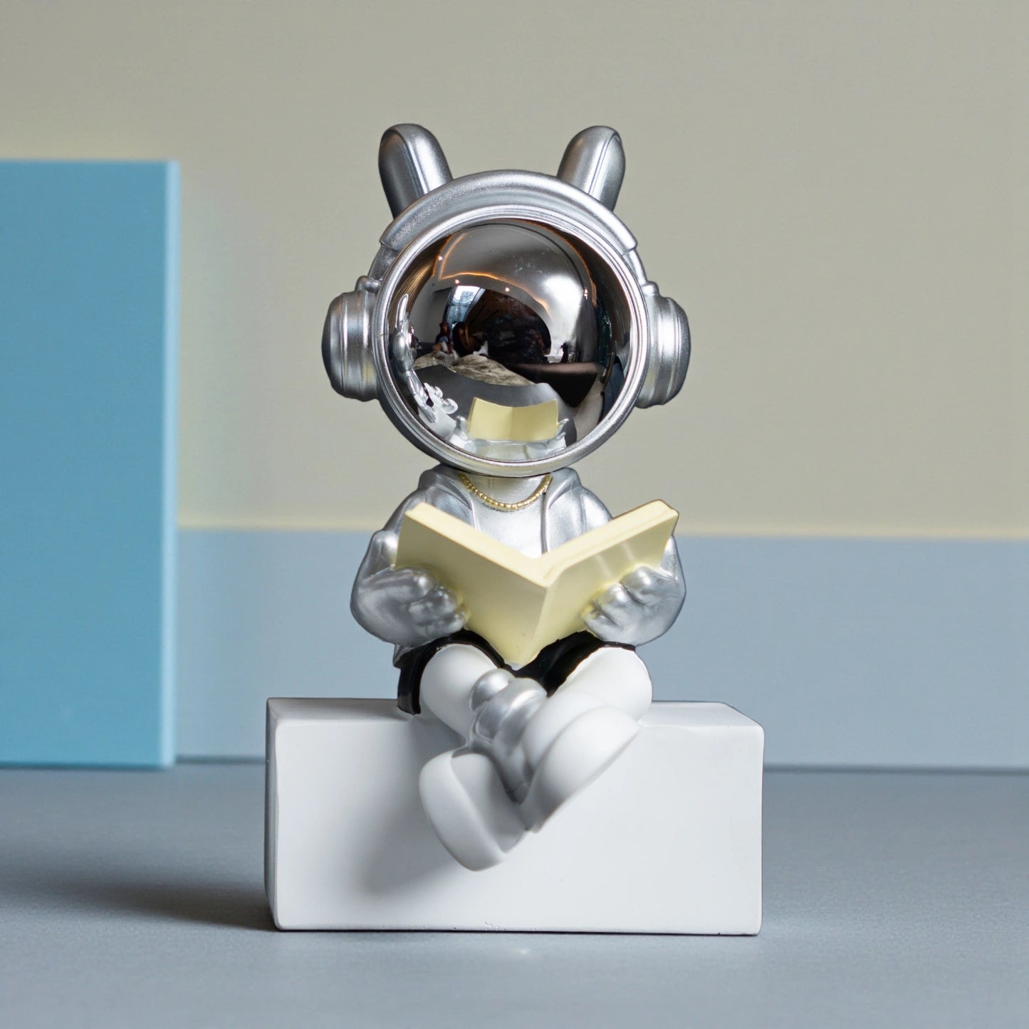 Astronaut figurine with chrome visor, silver suit, black shorts and cross-legged reading pose sitting on a white block; modern resin décor showpiece