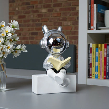 Astronaut figurine with chrome visor, silver suit, black shorts and cross-legged reading pose sitting on a white block; modern resin décor showpiece