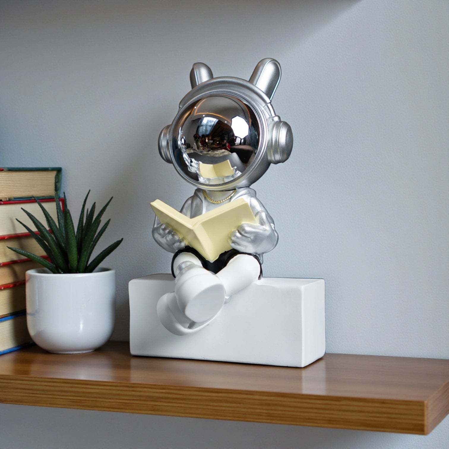 Astronaut figurine with chrome visor, silver suit, black shorts and cross-legged reading pose sitting on a white block; modern resin décor showpiece