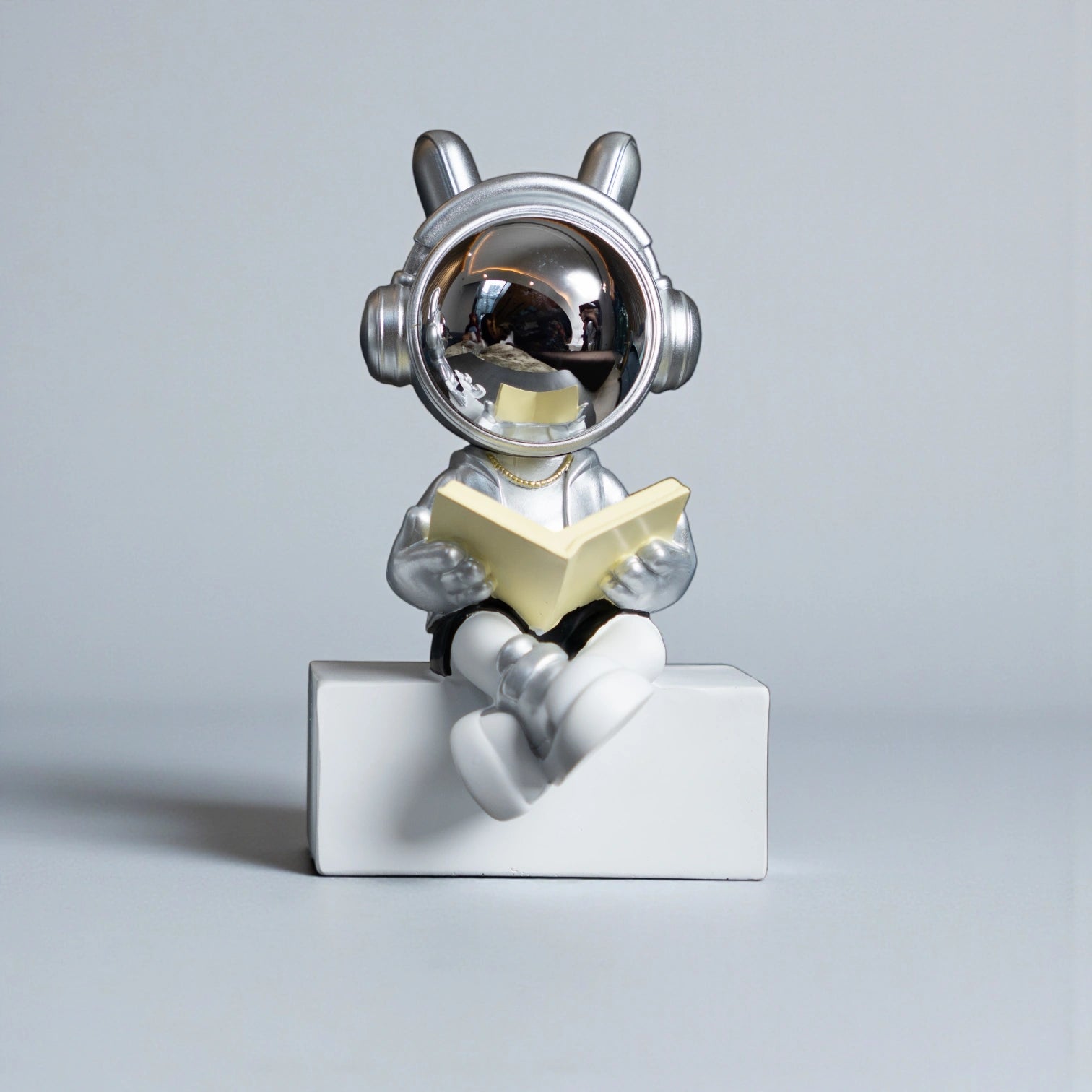 Astronaut figurine with chrome visor, silver suit, black shorts and cross-legged reading pose sitting on a white block; modern resin décor showpiece
