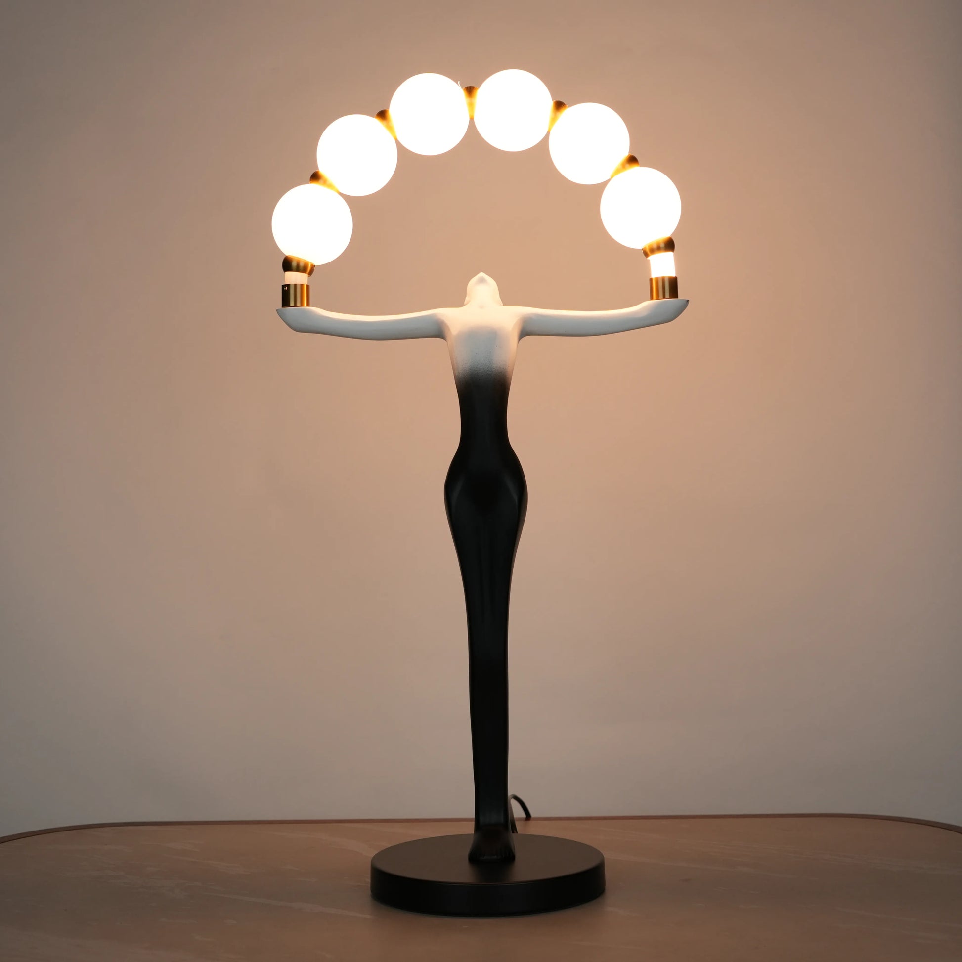 Small black-white ombre sculptural lamp featuring an elongated human figure lifting an arc of warm glowing spheres with gold accents on a round base.