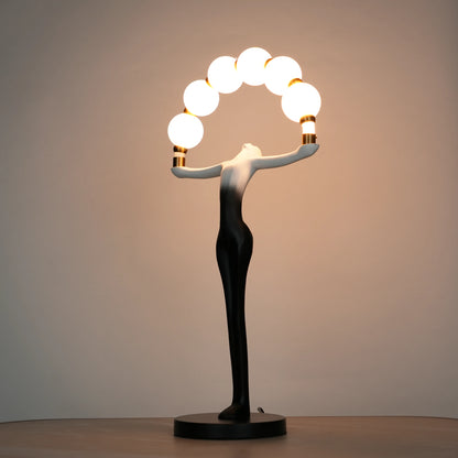 Small black-white ombre sculptural lamp featuring an elongated human figure lifting an arc of warm glowing spheres with gold accents on a round base.