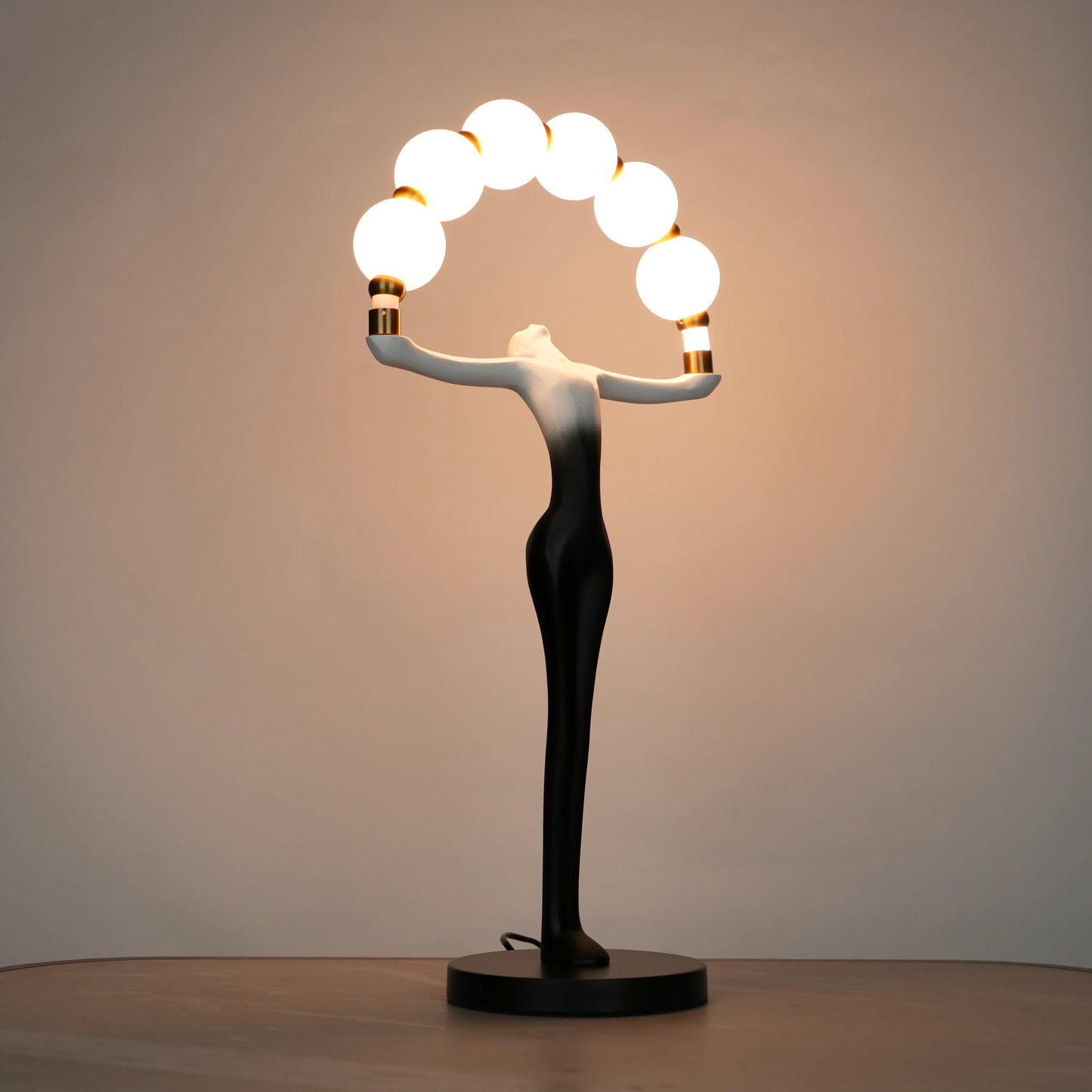 Small black-white ombre sculptural lamp featuring an elongated human figure lifting an arc of warm glowing spheres with gold accents on a round base.