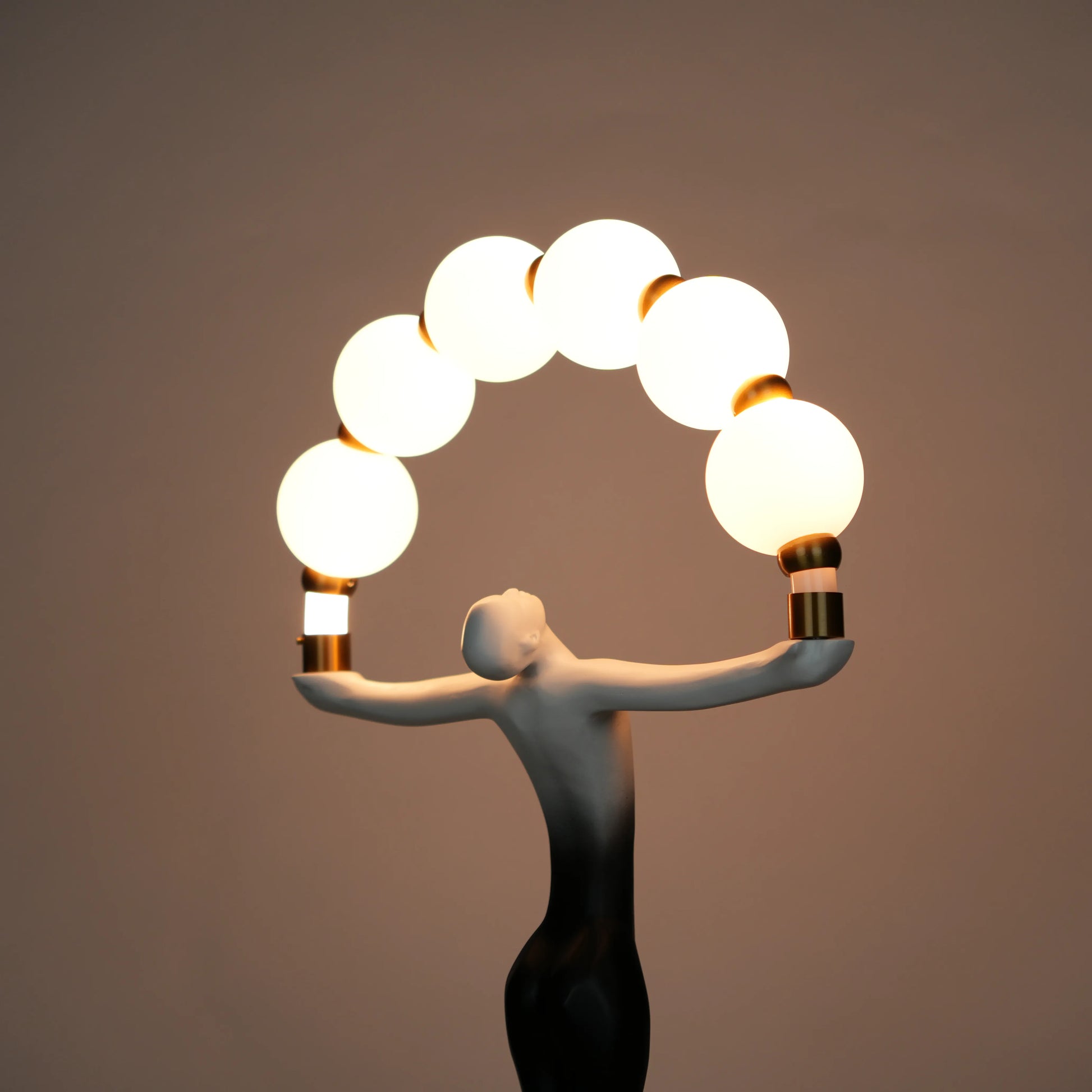 Small black-white ombre sculptural lamp featuring an elongated human figure lifting an arc of warm glowing spheres with gold accents on a round base.