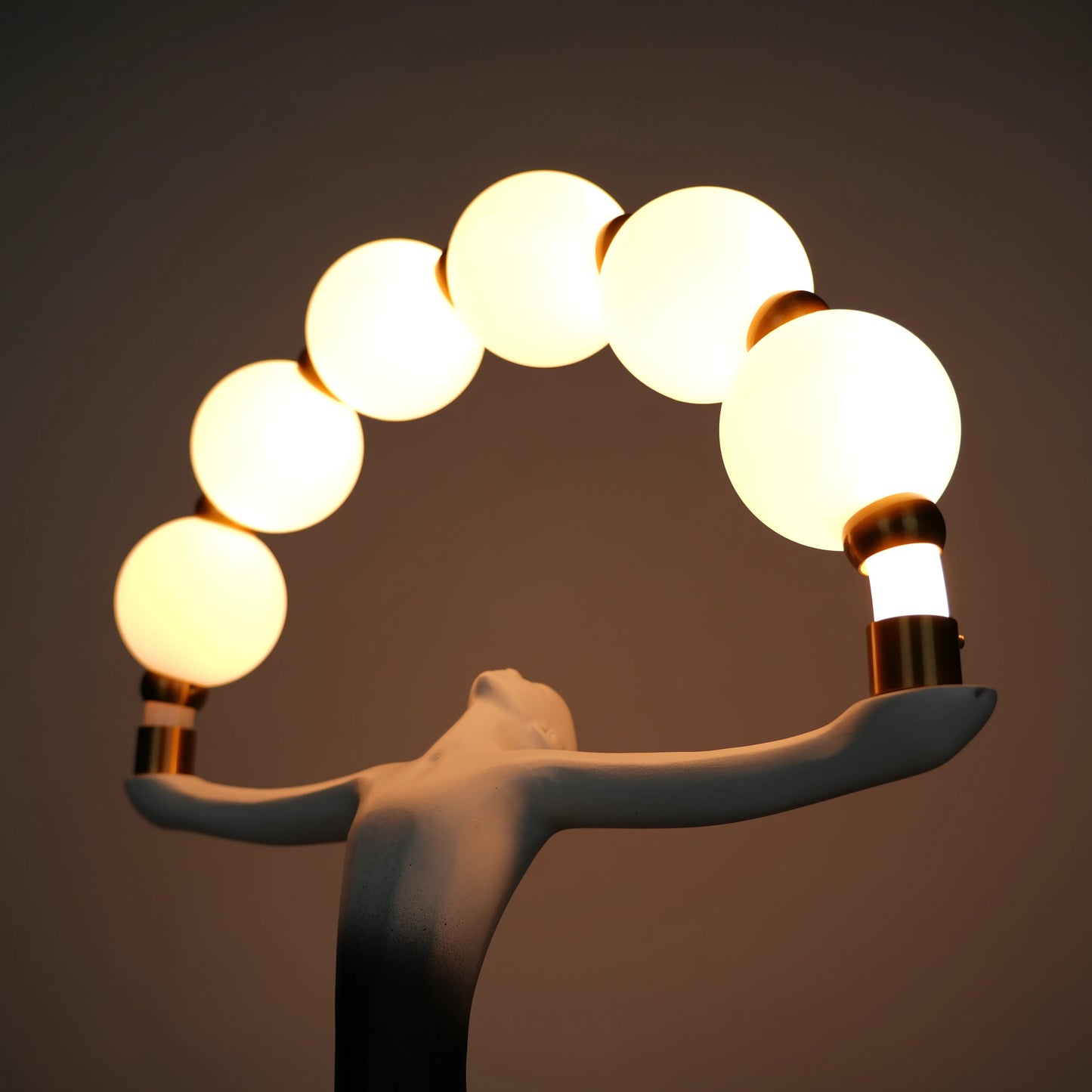 Small black-white ombre sculptural lamp featuring an elongated human figure lifting an arc of warm glowing spheres with gold accents on a round base.