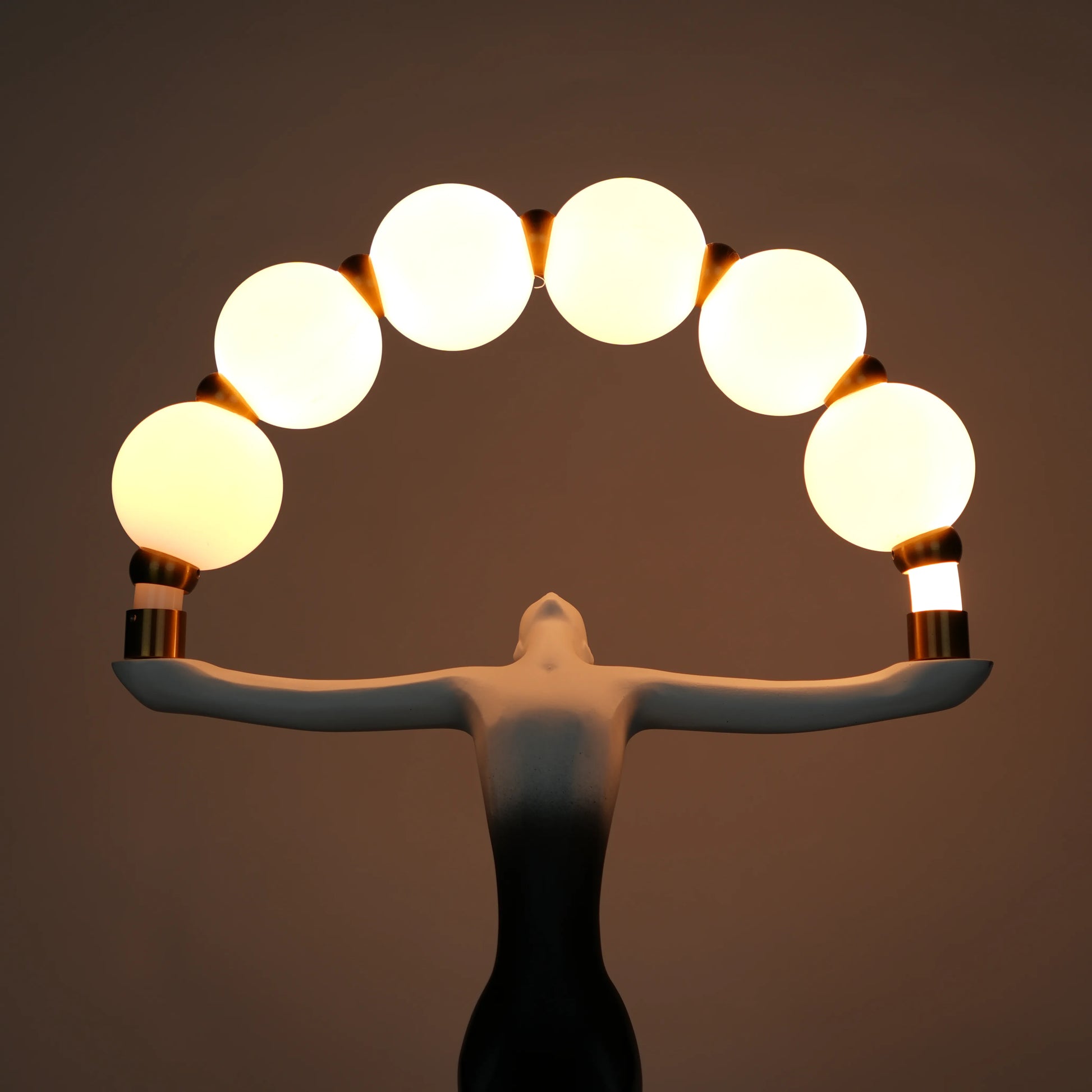 Small black-white ombre sculptural lamp featuring an elongated human figure lifting an arc of warm glowing spheres with gold accents on a round base.