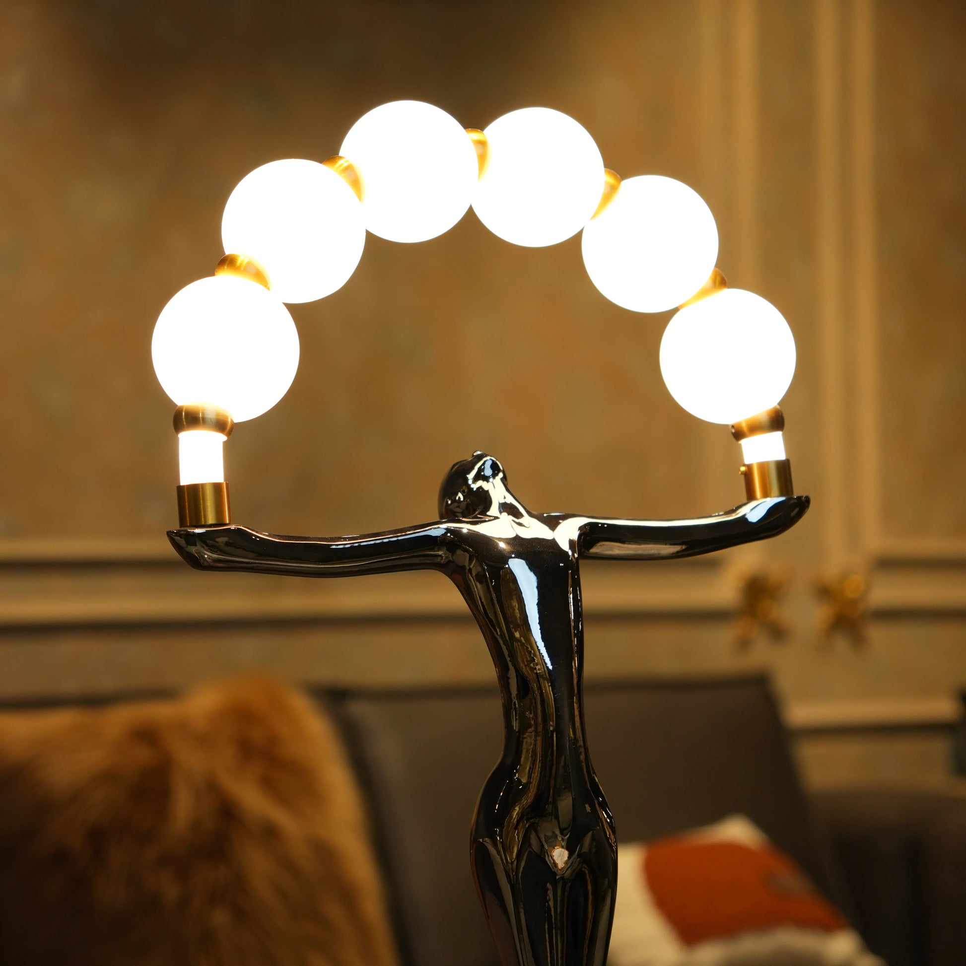 Small electroplated black human-form sculpture holding an arc of glowing warm orb lights with gold accents on a black circular base.