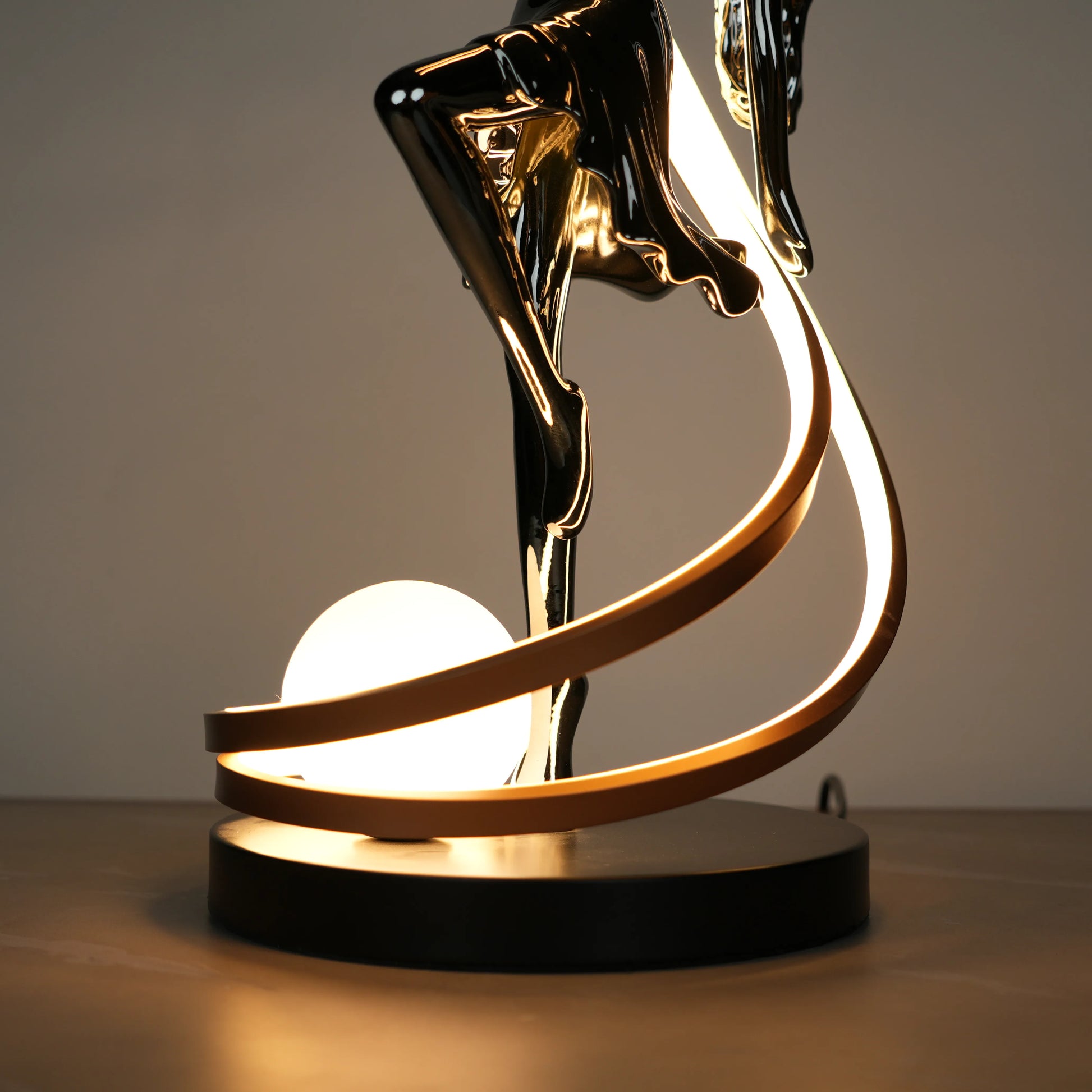 Small electroplated black sculpture lamp of a dancing woman with curved LED arcs and warm glowing orb.